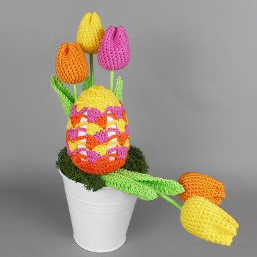 Gehäkeltes, bunt gemustertes Osterei mit fünf gehäkelten Tulpen in einem weißen Topf mit Moos