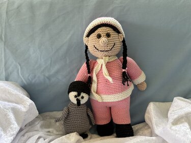 Gehäkelte Puppe in rosa mit geflochtenen schwarzen Zöpfen und kleinem grauem Amigurumi-Pinguin vor hellem Hintergrund