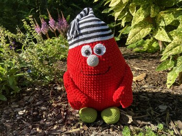 Rote, flauschige Amigurumi-Figur mit großen runden Augen, kleiner grauer Nase und schwarz-weiß gestreifter Mütze, sitzend im Garten.