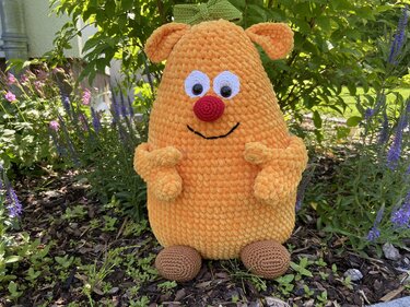 Orangefarbenes Amigurumi-Monster aus Chenillegarn mit roter Nase, großen Augen, grüner Schleife und braunen Füßen im Garten.
