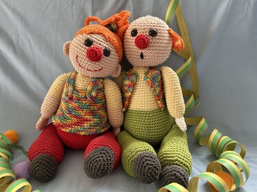 Zwei sitzende Amigurumi-Clown-Puppen mit roten Nasen, orangefarbenen Haaren und roter bzw. grüner Hose.