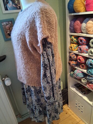 Schaufensterpuppe mit beigem, grobem Strickpullover und dunkelblauem Blumenkleid vor Regal mit Wolle