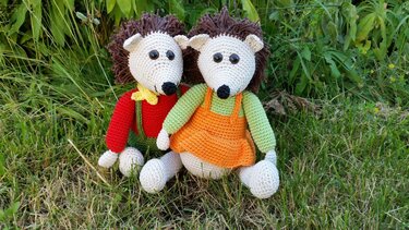 Zwei gehäkelte Igel-Amigurumi mit braunen Stacheln und bunten Kleidern sitzen im Gras