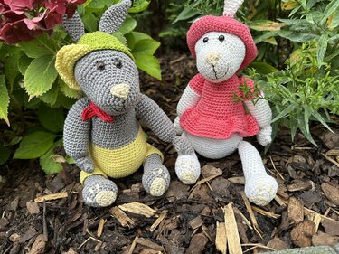 Amigurumi-Hasenpaar mit Outfits: grauer Hase mit gelber Hose, rotem Halstuch und grün-gelbem Hut, weißer Hase mit rosa Kleid und Hut.