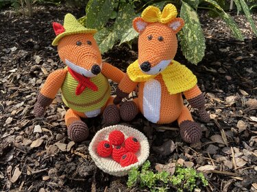 Nahaufnahme von zwei gehäkelten Fuchs-Amigurumi mit sichtbaren Maschen, Hut, Umhang und einer Schale mit roten Beeren