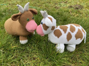 Zwei gehäkelte Amigurumi-Kühe mit Hörnern und pinken Schnauzen auf einer Wiese, eine braun und eine weiß mit braunen Flecken