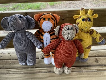 Vier gehäkelte Amigurumi-Tiere auf einer Holzbank: grauer Elefant, Tiger, Affe und gelbe Giraffe