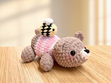 Gehäkelter brauner Bär mit rosa Backen und kleine gelb-schwarze Biene auf Holzoberfläche.