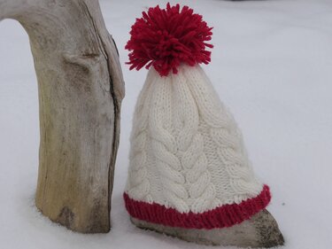 nahaufnahme weiße gestrickte Zopfmütze mit rotem geripptem Rand und rotem Bommel auf Schnee