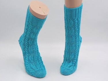 Zwei türkisfarbene, gestrickte Damensocken mit Zopf- und Ajourmuster auf Mannequin-Beinen vor weißem Hintergrund.