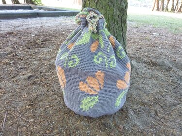 Gehäkelter Beutel in Grau mit orangefarbenen Blumen und grünen Blättern an einem Baumstamm