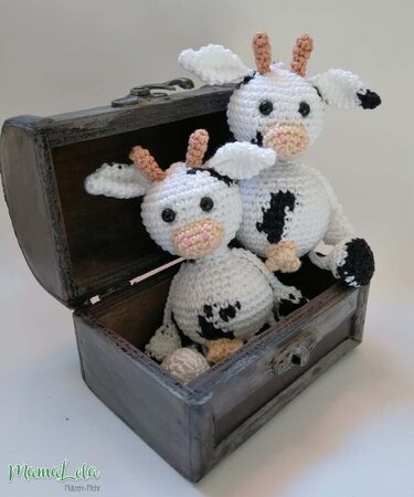 Zwei gehäkelte Kuh-Amigurumi in Weiß mit schwarzen Flecken und rosa Nüstern in einer offenen Holztruhe