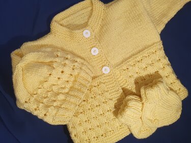 Ein gelbes gestricktes Baby-Set, bestehend aus Jäckchen, Mütze und Schühchen, auf dunkelblauem Hintergrund. Das Jäckchen hat weiße Knöpfe und ein feines Lochmuster.