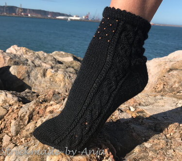 Schwarze gestrickte Damensocke mit Zopfmuster und Perlen am Bündchen vor Meereskulisse auf Felsen
