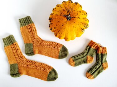 Gestrickte Damensocken und Babysocken in Orange und Oliv neben einem kleinen gelben Kürbis auf weißem Hintergrund