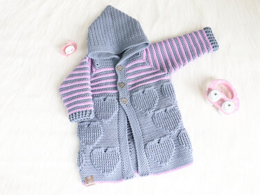 Grau-rosa gehäkelte Kapuzenjacke für Babys mit Streifen, Herztaschen und Herzknöpfen, liegend neben Schnuller und Spielzeug.