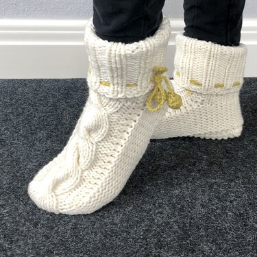 Zwei gestrickte cremefarbene Hüttenschuhe mit Zopfmuster und gelben Quasten auf dunklem Teppich