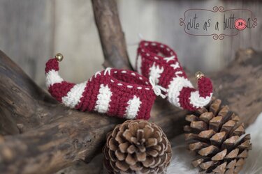 Zwei rot-weiß gestreifte, handgehäkelte Elfen-Babystiefel mit kleinen Glöckchen an den Spitzen, auf Holz neben Tannenzapfen arrangiert.