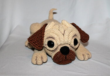 Beige and brown crocheted pug amigurumi lying down with large black eyes