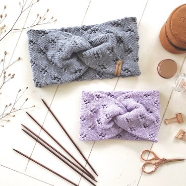 Zwei gestrickte Stirnbänder in Hellblau und Lavendel mit Lochmuster und gedrehter Vorderseite auf heller Holzoberfläche neben Stricknadeln und Zubehör.