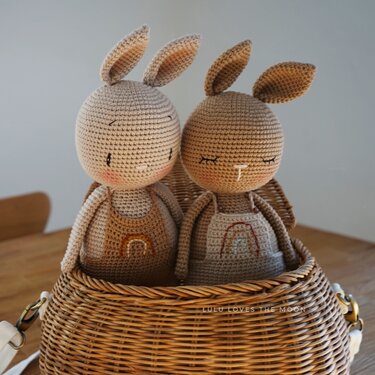 Zwei gehäkelte Amigurumi-Hasen mit Regenbogen-Overalls in einem Weidenkorb.