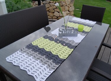 Gehäkelter Tischläufer mit Wellenmuster in Hellgrün, Grau und Weiß auf einem Glastisch im Garten, dekoriert mit Kerzen und einer Blüte.