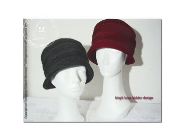 Zwei gehäkelte Damenhüte auf weißen Schaufensterköpfen, einer dunkelgrau, einer weinrot.