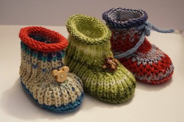 Drei gestrickte Babyschuhe nebeneinander: blau-beige, grün mit Knopf, rot-blau meliert