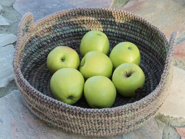 Handmade round crocheted basket with two handles holding seven green apples on a stone surface