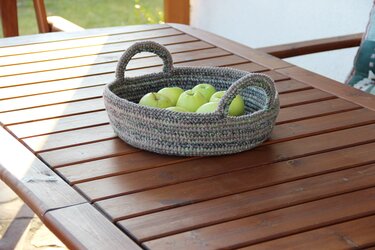 Round crocheted basket in grey, brown and green yarn holding green apples on a wooden table.