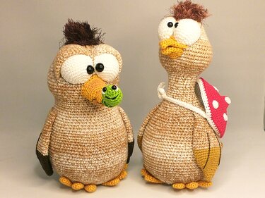 Two tan crocheted amigurumi birds; left holds a small green crochet frog in its beak, right wears a red polka-dot mushroom cap backpack
