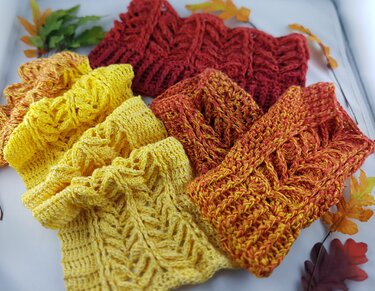 Drei handgehäkelte Stulpen in Gelb, Orange und Bordeauxrot mit Zopfmustern, dekoriert mit Herbstblättern.