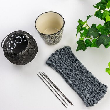 Textured gray knitted headband, three double-pointed knitting needles, patterned ceramic mug, ornamental metal lantern, and green plant on a white background.