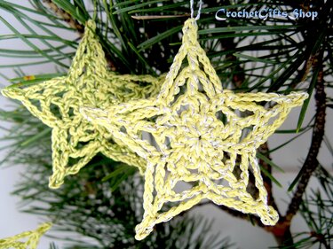 Two pale yellow crocheted star ornaments with silver metallic thread hanging on pine needles.