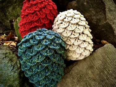 Drei gehäkelte Dekoobjekte mit Schuppenmuster in Rot, Creme und Türkis auf Steinen