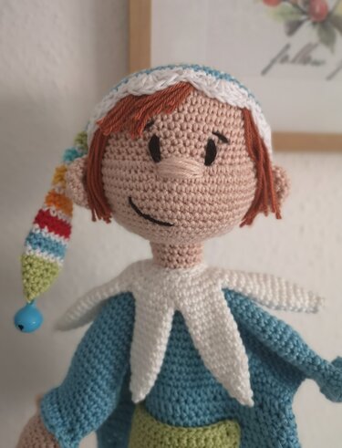 Gehäkelte Amigurumi-Puppe mit orangebraunem Haar, blauem Oberteil und gestreifter Zipfelmütze mit blauer Perle