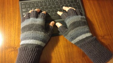 Hände mit gestreiften, fingerlosen Strickhandschuhen tippen auf einer schwarzen Tastatur auf einem Holztisch.