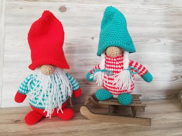Zwei gehäkelte Weihnachtswichtel mit roten und türkisen Mützen, weißem Bart und Zöpfen, einer auf einem Holzschlitten