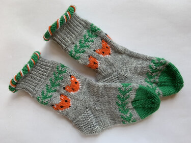 Ein Paar grauer gestrickter Babysocken mit orangefarbenen Fuchsgesichtern, grünen Zehen und einem gedrehten Bündchen.