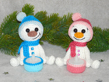 Two crocheted amigurumi snowmen wearing blue and pink hats with matching crocheted tealight holders and pine sprig background.