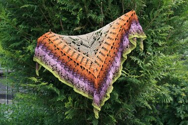 Gehäkeltes Dreieckstuch mit Farbverlauf in Beige, Orange, Lila und Grün, an einer immergrünen Hecke drapiert.