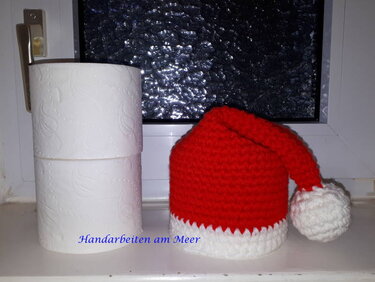 Kleine rote gehäkelte Weihnachtsmütze mit weißem Rand und Pompon auf einer Fensterbank neben zwei Toilettenpapierrollen.
