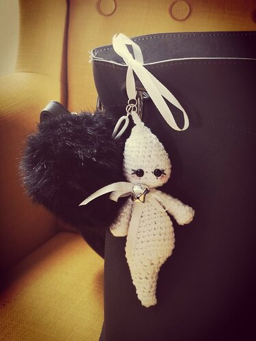 Weißer gehäkelter Geist-Schlüsselanhänger mit Glöckchen an dunkler Handtasche neben schwarzem Pompon