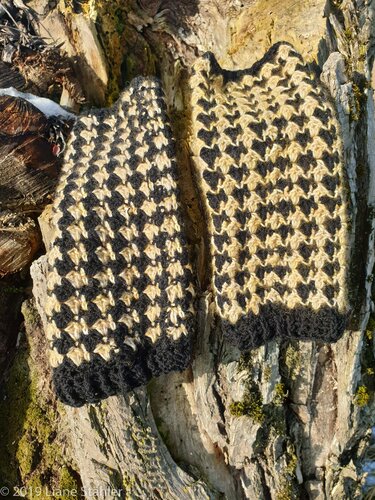 Beige‑schwarze gehäkelte Stulpen mit Hahnentritt‑ähnlichem Muster, auf Rinde arrangiert