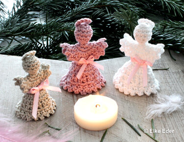 Drei kleine DIY-Häkelengel in beige, rosa und weiß mit rosa Schleifen neben einem Teelicht
