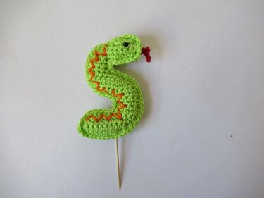 Hellgrüne gehäkelte Schlange mit orangefarbenem Zickzackmuster und Holzspieß auf weißem Hintergrund