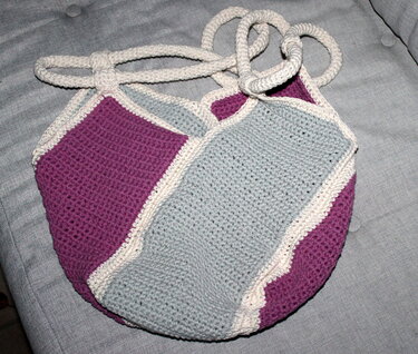 Gehäkelte Shopper-Tasche mit lila und hellblauen Farbblöcken, cremefarbener Kante und gehäkelten Tragegriffen auf grauem Sofabezug.