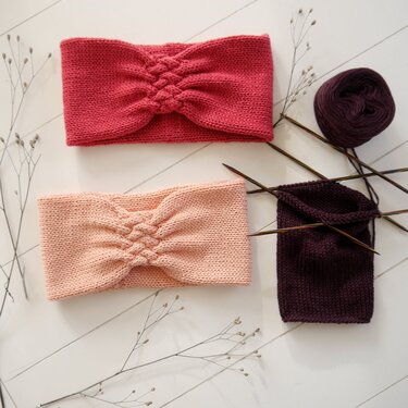 Zwei gestrickte Stirnbänder in Rosa und Koralle mit Zopfmuster neben einem angefangenen dunkelvioletten Strickstück, Stricknadeln und einem Wollknäuel.