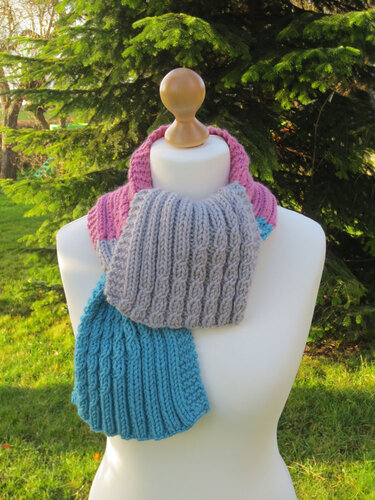 Dreifarbiger gestrickter Schal (Pink, Grau, Türkis) mit Zopf- und Rippenmuster auf weißer Schaufensterpuppe vor Bäumen
