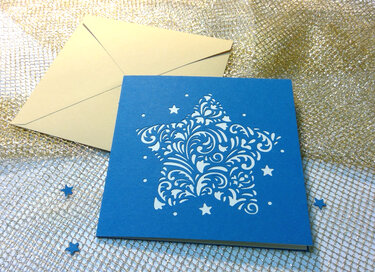 Blaue Karte mit großem, verziertem Sternausschnitt und beigefarbenem Umschlag auf goldenem Netzstoff.